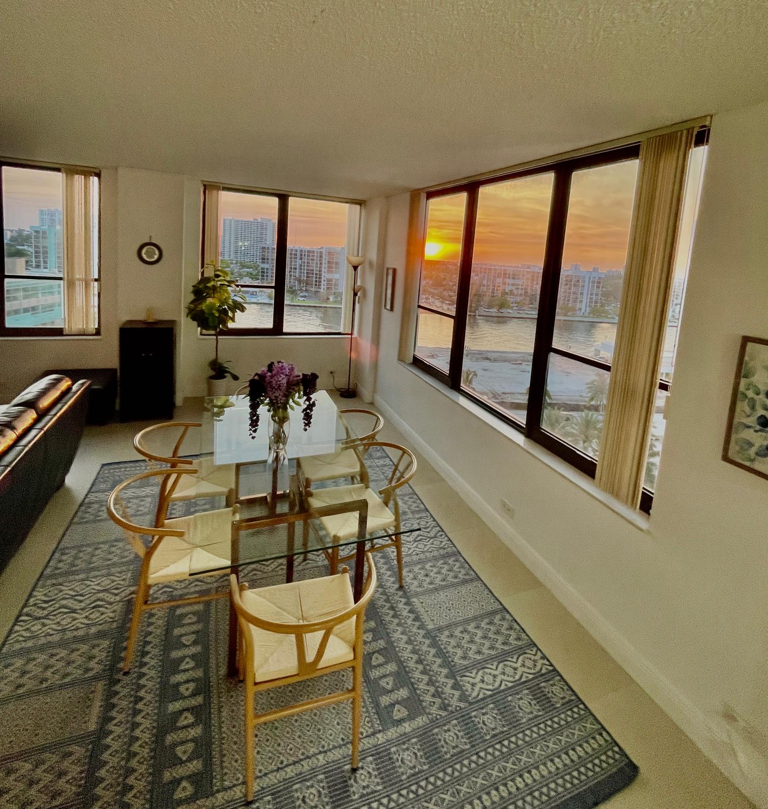Glass dining table with six wishbone chairs beside corner windows glowing at sunset