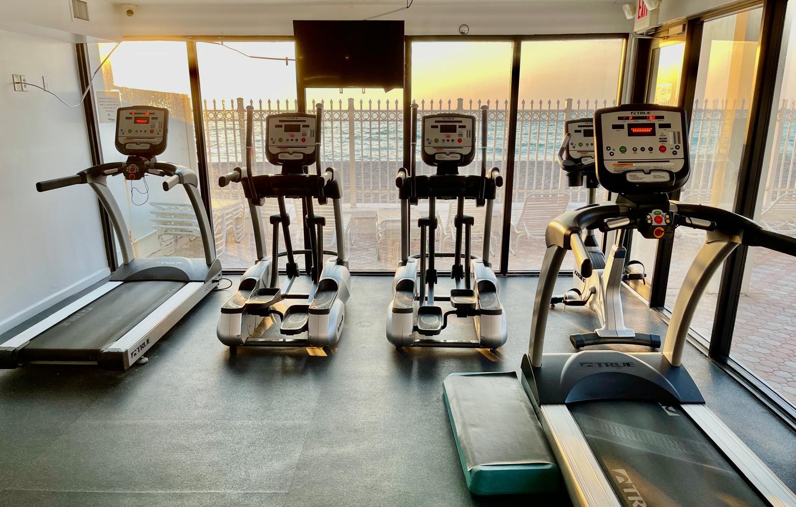 Treadmills and ellipticals in the fitness room facing floor-to-ceiling windows onto the beach
